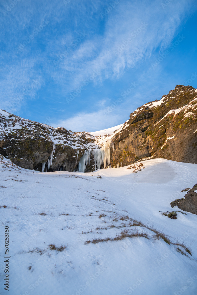 Fully snow covered mountain towards the Kvernufoss waterfall and a gorge with snow covered areas and stalagmites illuminated with the early morning sun illuminating the snow capped peaks of Iceland