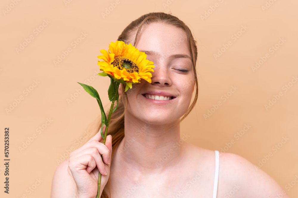 Young caucasian girl isolated on beige background holding a sunflower while smiling. Close up portrait