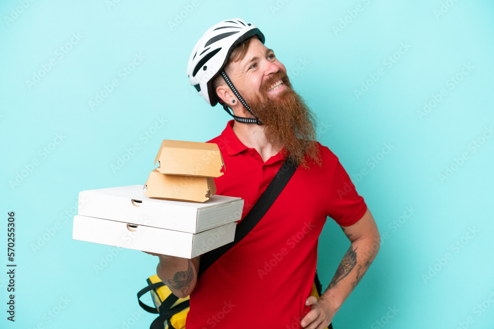 Delivery man holding pizzas and burgers isolated on blue background posing with arms at hip and smiling Delivery man holding pizzas and burgers isolated on blue background posing with arms at hip and smiling