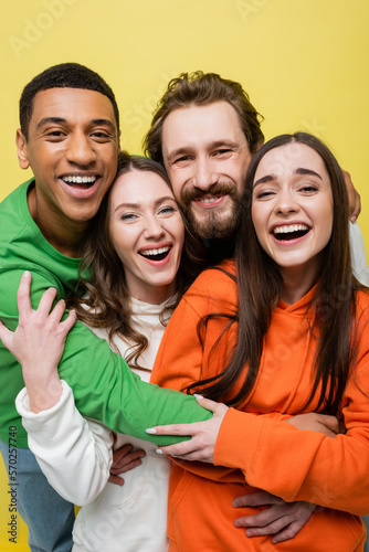 Happy interracial friends hugging and looking at camera isolated on yellow