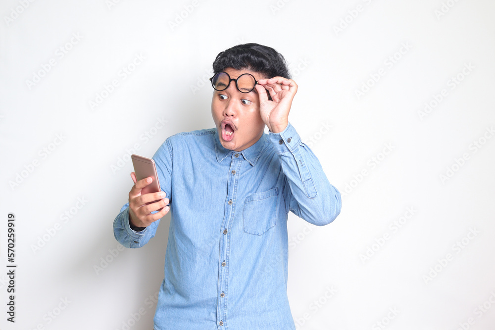Portrait of shocked Asian man in blue shirt looking at mobile phone. Wow face expression. Isolated image on white background