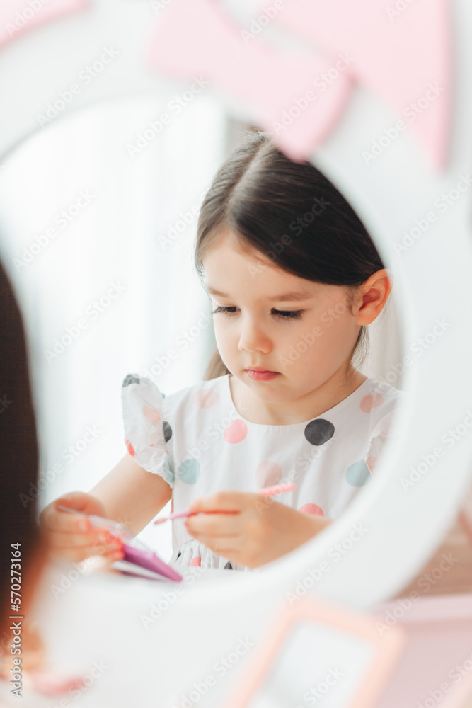 a beautiful little girl in the mirror preening. a little girl is sitting at a children's table and is wearing children's cosmetics. a beautiful little girl in the mirror preening. a little girl is sitting at a children's table and is wearing children's cosmetics.