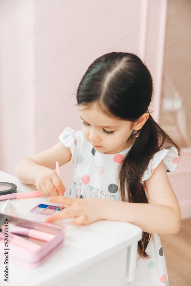 a beautiful little girl in the mirror preening. a little girl is sitting at a children's table and is wearing children's cosmetics. a beautiful little girl in the mirror preening. a little girl is sitting at a children's table and is wearing children's cosmetics.