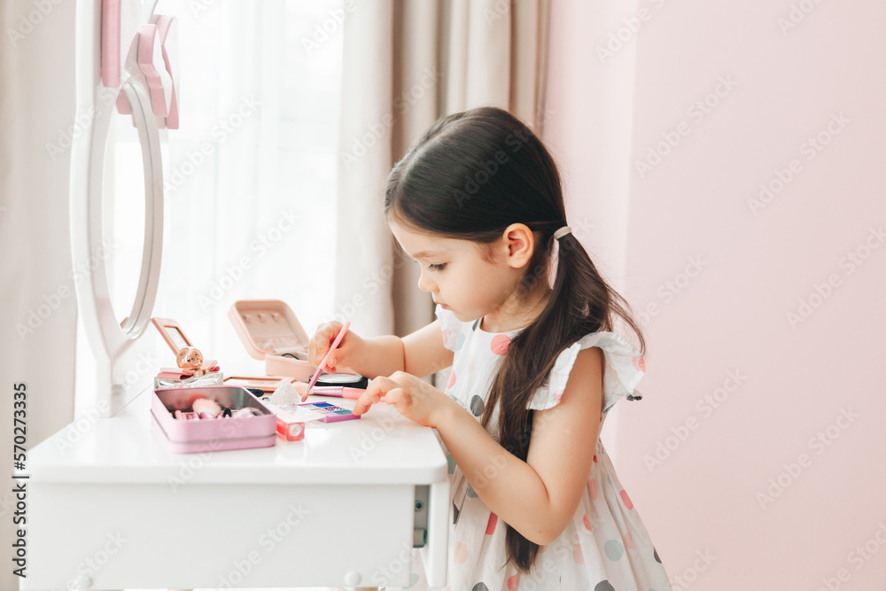 a beautiful little girl in the mirror preening. a little girl is sitting at a children's table and is wearing children's cosmetics. a beautiful little girl in the mirror preening. a little girl is sitting at a children's table and is wearing children's cosmetics.