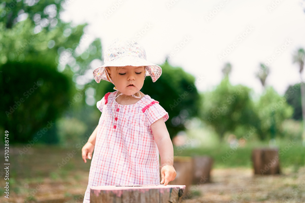 Little girl in a panama hat stands on the lawn near the stump