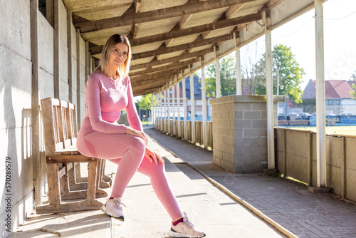 Street Workout. Pretty blonde lady in pink sports clothes resting after training on sunny day, sitting on amateur soccer tribune bench, taking care of her body. High quality photo