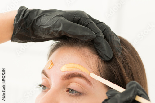 Young woman undergoing eyebrow correction procedure, waxing, wax depilation of eyebrow hair, brow correction in beauty salon, closeup. Golden wax and wooden sticks, tools. Eyebrow master