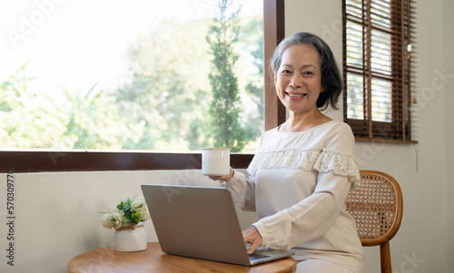 Happy aged Asian old woman working , sipping coffee while using laptop