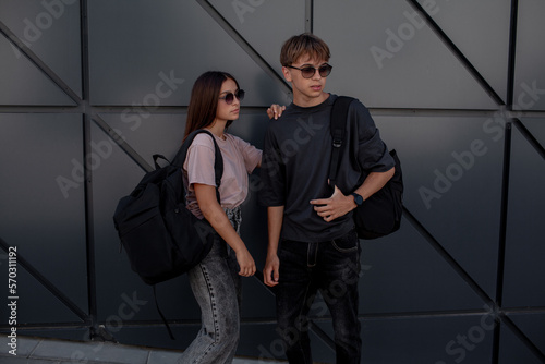 The fashion couple of girl and boy teenagers dressed black t-shirt, jeans, sunglasses, watches and bagback walking in the street at the background grey wall