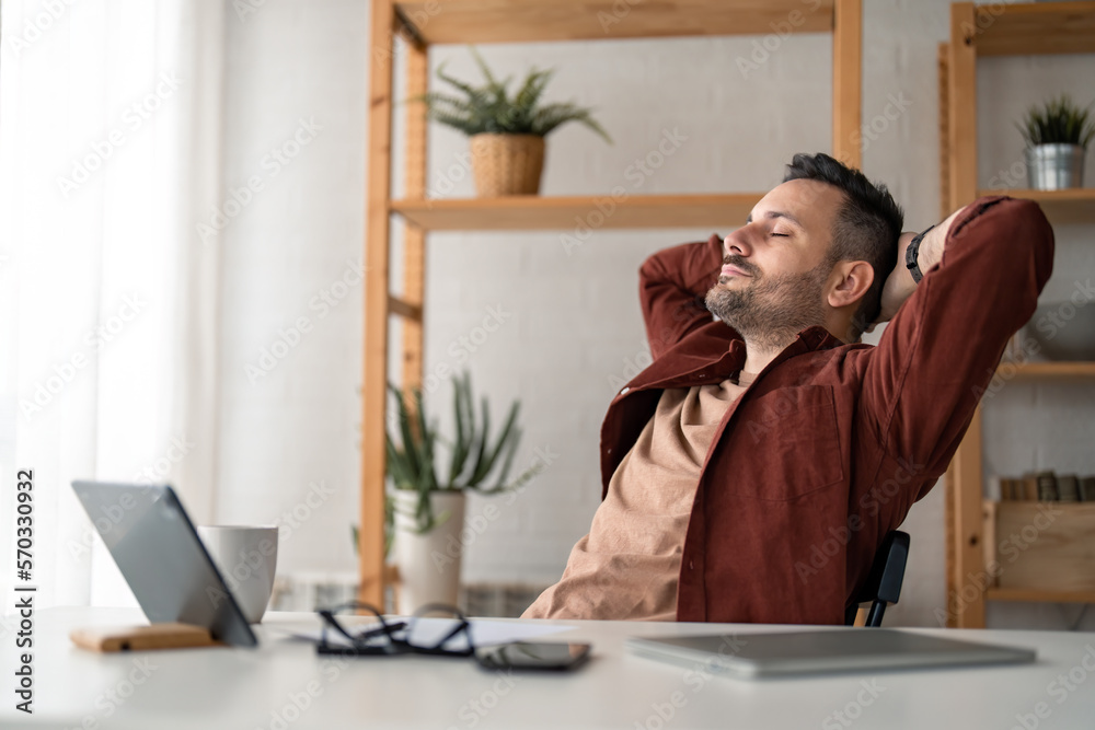Fototapeta premium Caucasian millennial business man manager resting after hard work in home office during the day with hands behind the back of the head, looking carefree and relaxed.
