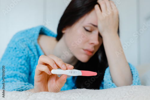 Pregnant test.Sad single woman complaining about holding a pregnancy test sitting on a couch in the living room at home covering eyes with hand
