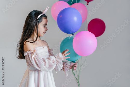 Pretty little brunette girl in a pink dress and crown with blue, green and pink balloons.