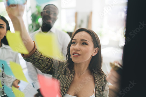 Positive female putting sticker on glass wall while standing with diverse colleagues