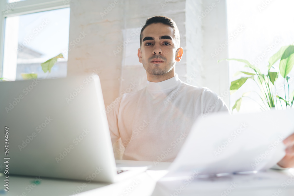 Focused young man with document in hand sitting at table with laptop in office Focused young man with document in hand sitting at table with laptop in office