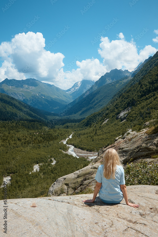 a girl sits on the edge of a cliff and looks into the distance, in the distance there is a river and a gorge, a good mood, clouds and a mountain landscape