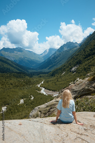 a girl sits on the edge of a cliff and looks into the distance, in the distance there is a river and a gorge, a good mood, clouds and a mountain landscape