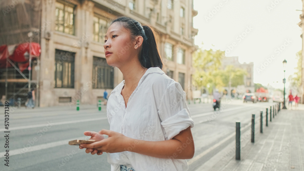 Young woman using mobile phone while standing next to the road. Girl looks around