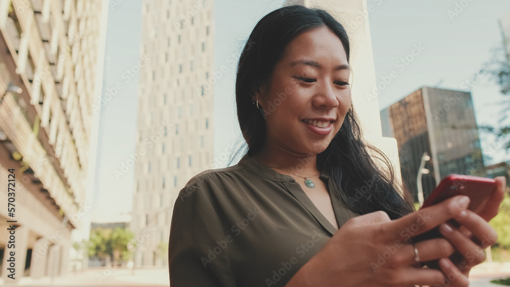 Fototapeta premium Close-up of young happy businesswoman using mobile phone while standing on modern buildings background