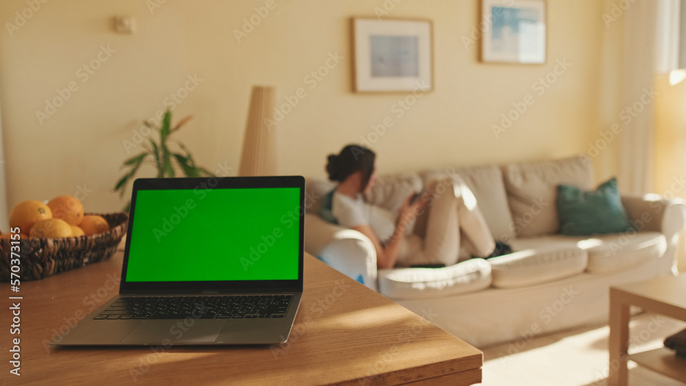 Foreground on the table is laptop with green screen chroma key, in the background girl is sitting, soft focus Foreground on the table is laptop with green screen chroma key, in the background girl is sitting, soft focus