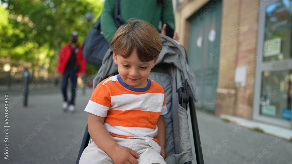 Little boy seated on stroller during beautiful sunny day in city street. Closeup face of child strolling outdoors with parent Little boy seated on stroller during beautiful sunny day in city street. Closeup face of child strolling outdoors with parent