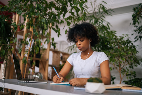Focused African-American female student wearing headphones writing notes, watching webinar, studying online using laptop, sitting at table in green home office room with modern biophilia design.