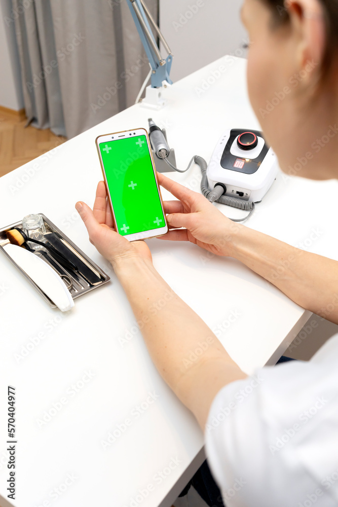 Manicure, pedicure technician holds smartphone with green chroma key screen, sitting at workstation in nail beauty treatment salon. Professional tools are on white table. Studying manicurist. Vertical