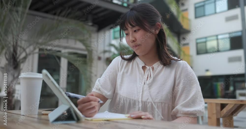 Young asian girl with freckles thinking and studying online with a tablet, writing notes with a pen outdoor cafe in slow motion Young asian girl with freckles thinking and studying online with a tablet, writing notes with a pen outdoor cafe in slow motion