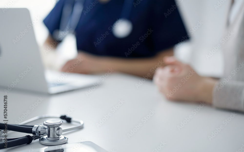Stethoscope lying on the tablet computer in front of a doctor and patient using laptop computer at the background. Medicine, healthcare concept Stethoscope lying on the tablet computer in front of a doctor and patient using laptop computer at the background. Medicine, healthcare concept