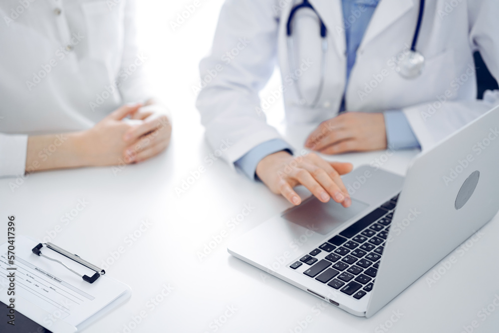 Doctor and patient sitting near each other at the desk in clinic. The focus is on female physician's hands using laptop computer, close up. Medicine concept Doctor and patient sitting near each other at the desk in clinic. The focus is on female physician's hands using laptop computer, close up. Medicine concept
