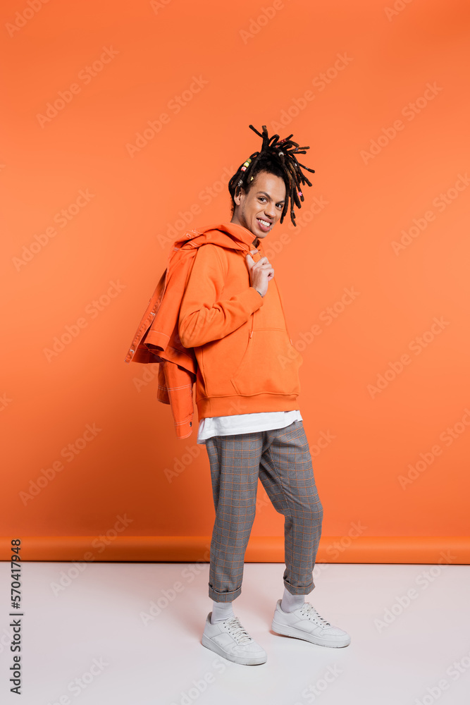Fototapeta premium full length of positive multiracial man in hoodie holding denim jacket on coral background