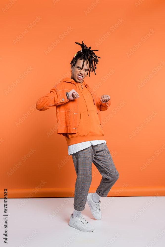 Fototapeta premium full length of excited and multiracial man with dreadlocks standing with clenched fists on coral background
