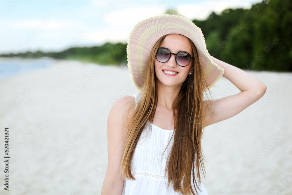 Portrait of a happy smiling woman in free happiness bliss on ocean beach standing with a hat and sunglasses. A female model in a white summer dress enjoying nature 
