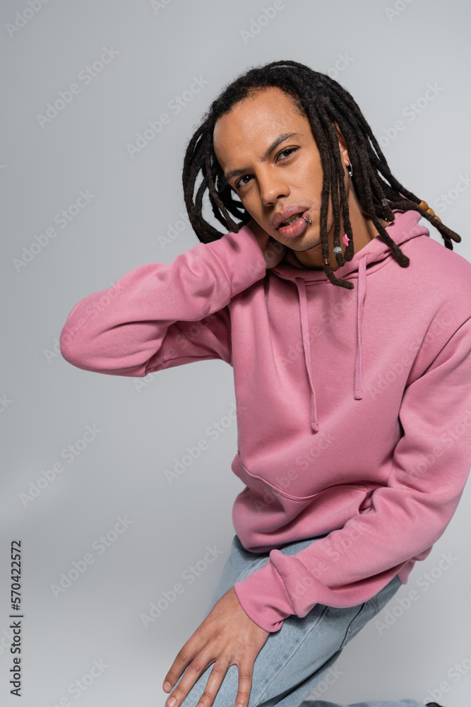 Fototapeta premium pierced multiracial man with dreadlocks looking at camera and posing on grey