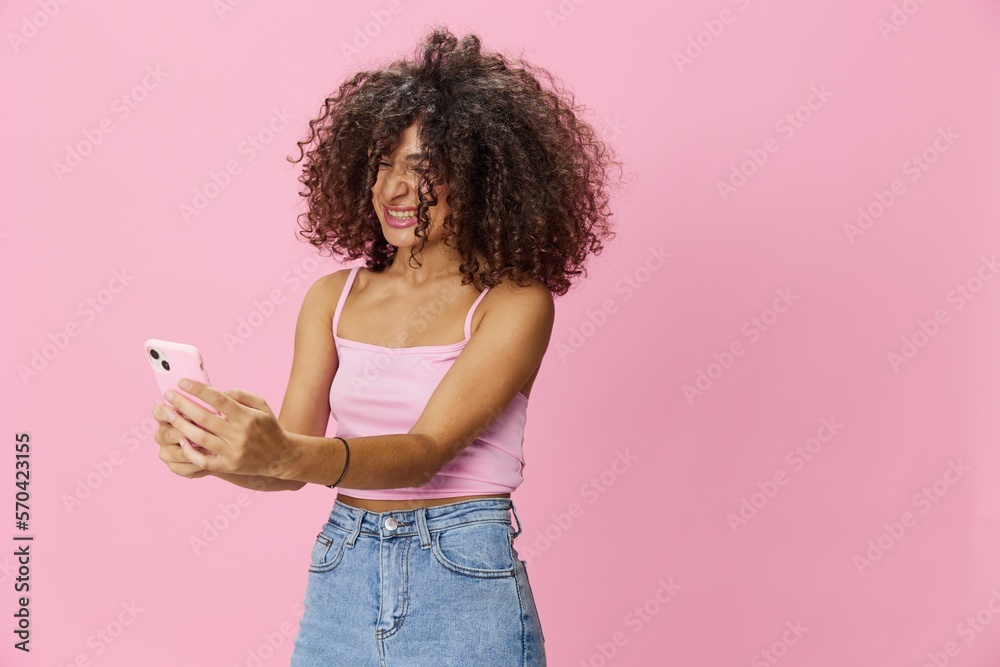 Woman blogger holding phone in hand video call, with curly hair in pink top and jeans poses on pink background, copy space, technology and social media, online Woman blogger holding phone in hand video call, with curly hair in pink top and jeans poses on pink background, copy space, technology and social media, online