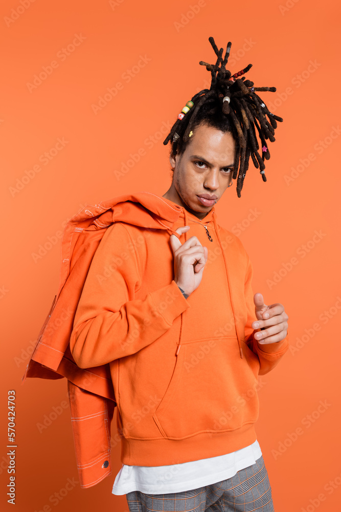tattooed and multiracial man in hoodie holding denim jacket isolated on coral background tattooed and multiracial man in hoodie holding denim jacket isolated on coral background