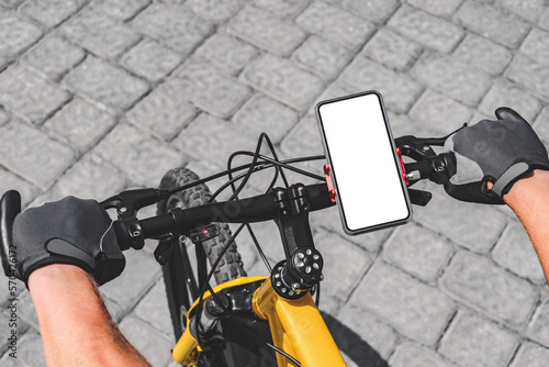 Fototapeta Naklejka Na Ścianę i Meble -  Mockup of a smartphone on the handlebars of a bicycle with an action camera, with the hand of a person with red gloves in the city. on the background of the road with tiles.