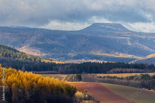 A view of the Szczeliniec Mountain Peak, towering over the Kłodzko Valley. Autumn trees illuminated by the sun.