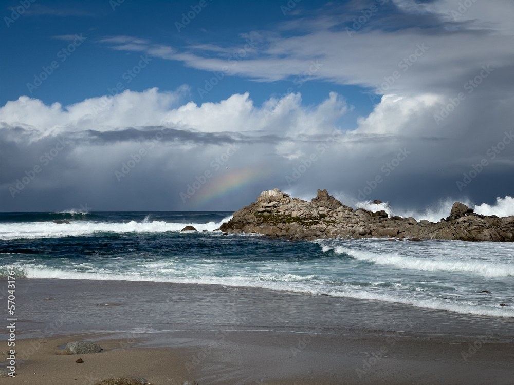 Rainbow on horizon with storm clouds in the sky with waves crashing off shore on rock formation at beach in Monterey County, California
