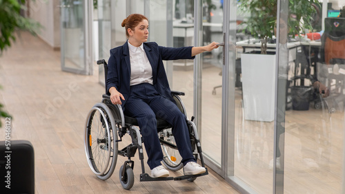 Red-haired caucasian woman in a wheelchair trying to open the door in the office.  © Михаил Решетников