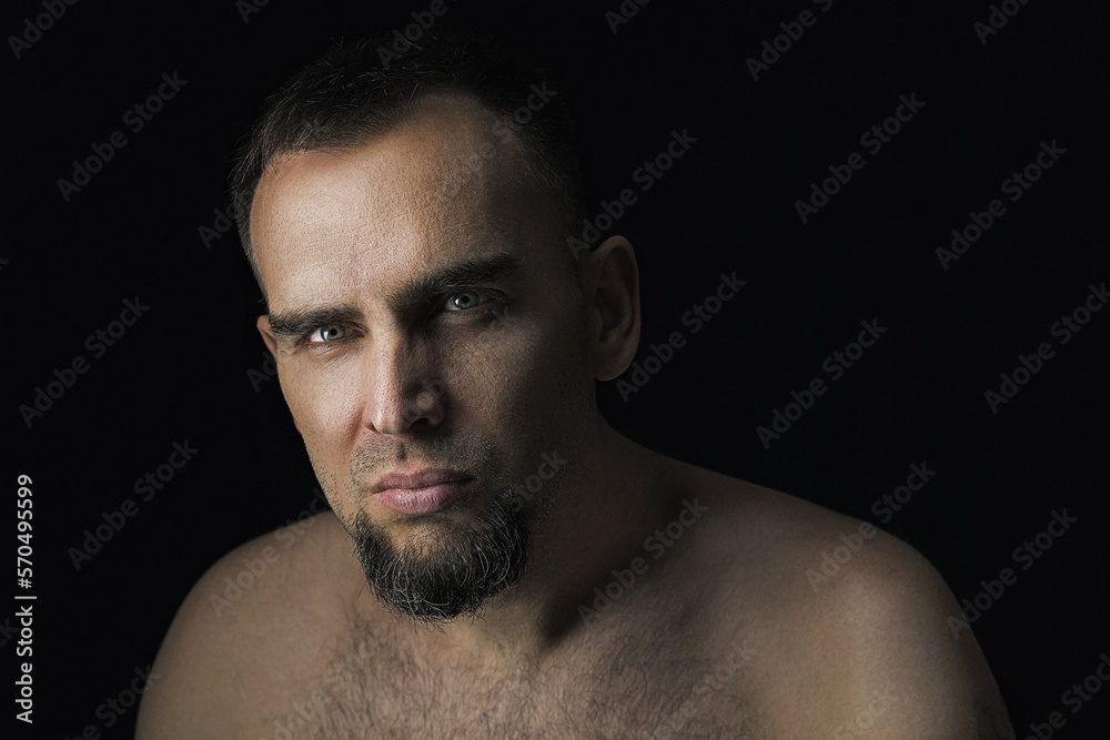 A dramatic portrait of a handsome man in an unobtrusive setting. Close up of a handsome bearded man with no clothes on a black background. Sexy man looking into the camera.