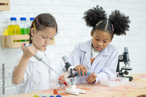 Two little kids in lab coat learning chemistry in school laboratory. Young scientists in protective glasses making experiment in lab or chemical cabinet. Working on a tablet.