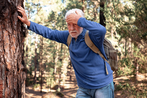 Senior bearded man feeling bad while walking in the mountain woods. Elderly man with backpack touching his head leaning against a tree trunk to catch his breath