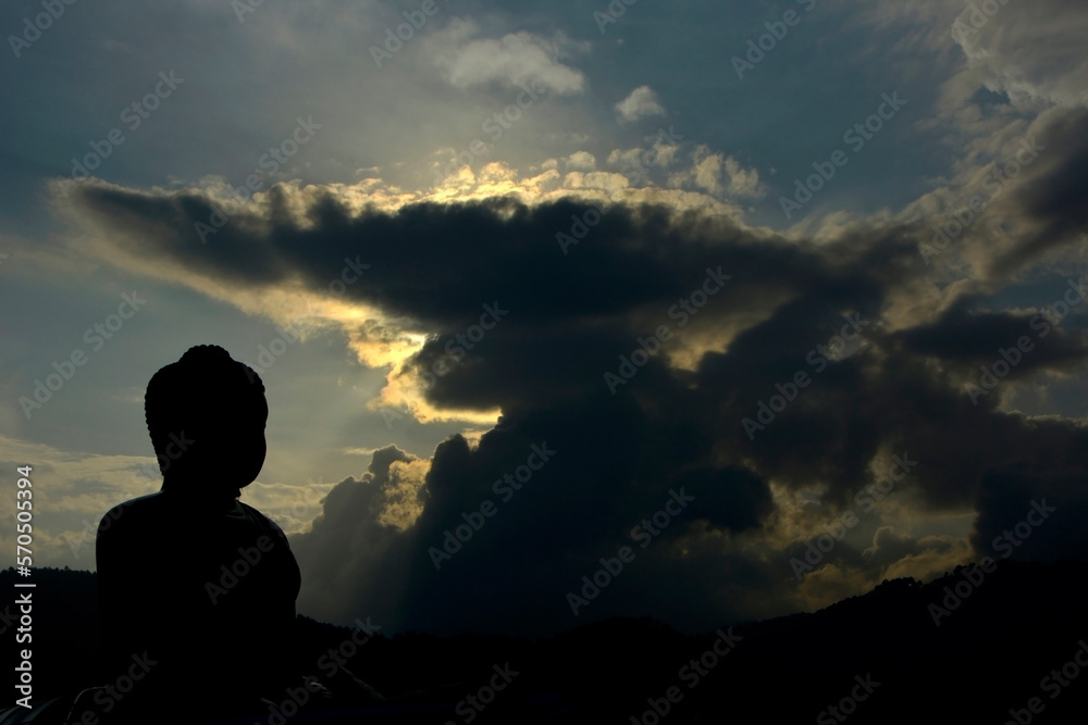 A shadowy silhouette of a Buddhist black stone Buddha in nature with a huge black cloud in the background.