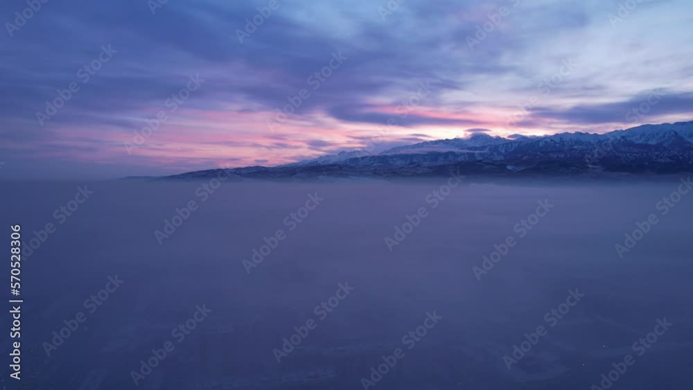 Dawn over the city in fog and smog. Mountain view. A light haze hangs over the city, cars drive, signs glow. Clouds, sun rays and snowy mountains are visible in the distance. Almaty, Kazakhstan Dawn over the city in fog and smog. Mountain view. A light haze hangs over the city, cars drive, signs glow. Clouds, sun rays and snowy mountains are visible in the distance. Almaty, Kazakhstan