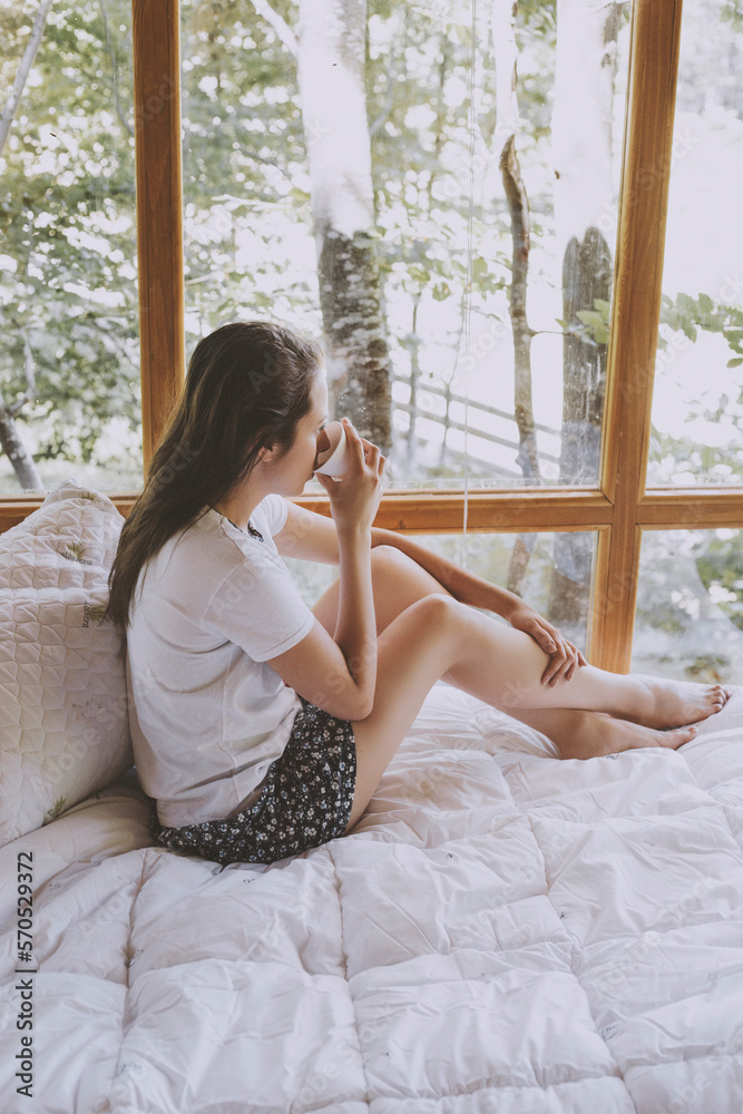 Young woman sitting on bed, drinking coffee and looking through window Young woman sitting on bed, drinking coffee and looking through window