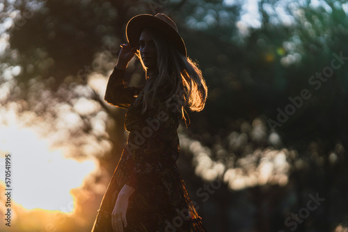 Chica joven delgada de pelo largo con sombrero y ropa otoñal posando al atardecer en un bosque