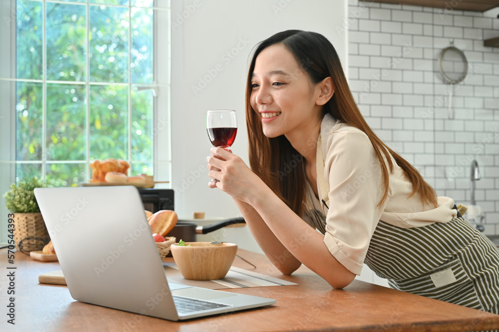 Pretty young woman drinking red wine and using laptop in modern kitchen. People, technology and lifestyle concept Pretty young woman drinking red wine and using laptop in modern kitchen. People, technology and lifestyle concept