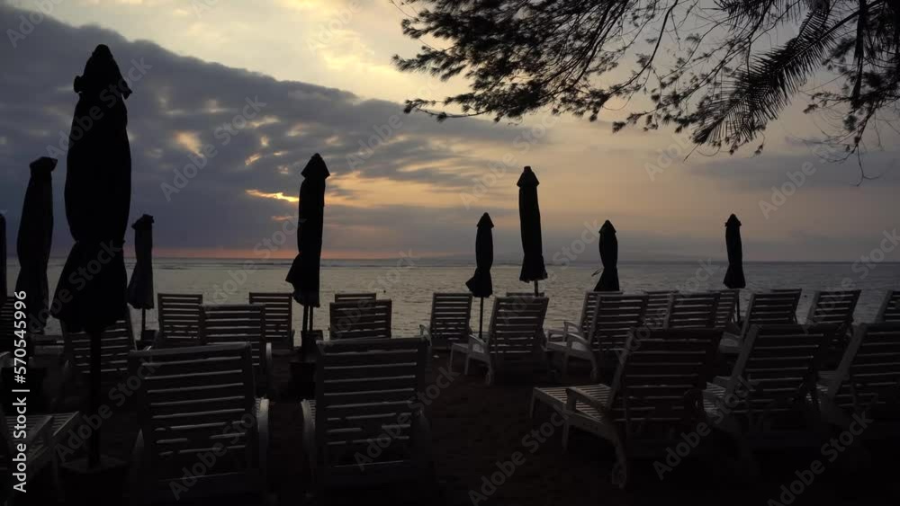 Dolly shot of silhouette deck chairs with parasols arranged on Sanur Beach against cloudy sky during sunset