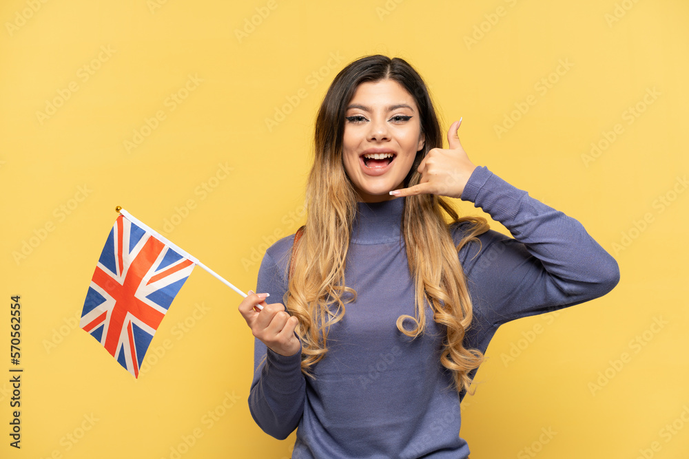 Young Russian girl holding an United Kingdom flag isolated on yellow background making phone gesture. Call me back sign Young Russian girl holding an United Kingdom flag isolated on yellow background making phone gesture. Call me back sign