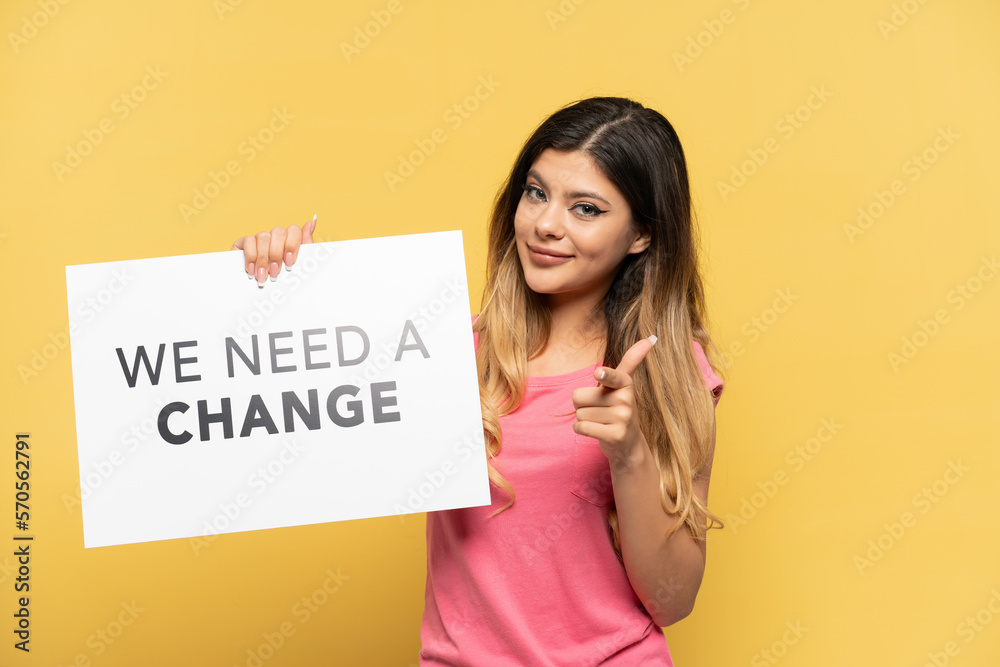 Young Russian girl isolated on yellow background holding a placard with text We Need a Change and pointing to the front Young Russian girl isolated on yellow background holding a placard with text We Need a Change and pointing to the front